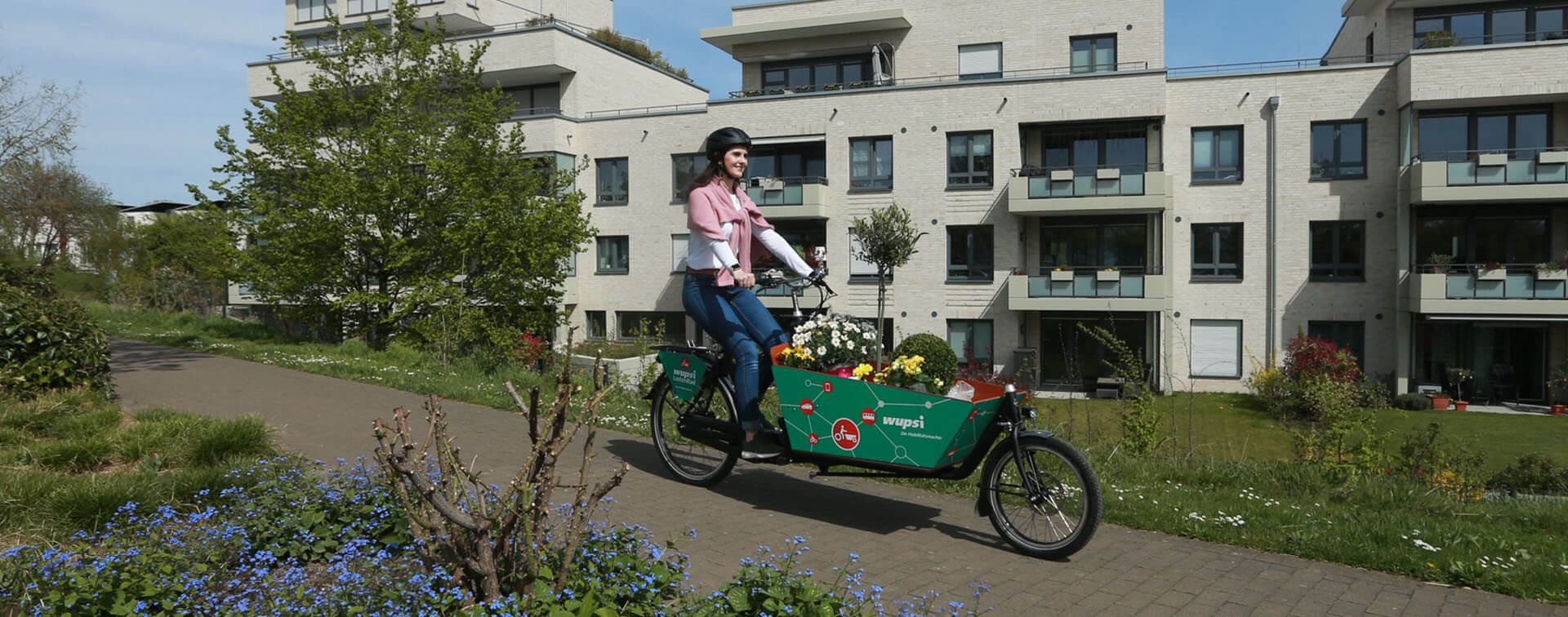 Eine Frau fährt glücklich mit ihrem Wupsilastenrad, in dem sie Blumen transportiert, durch eine Wohnsiedlung.